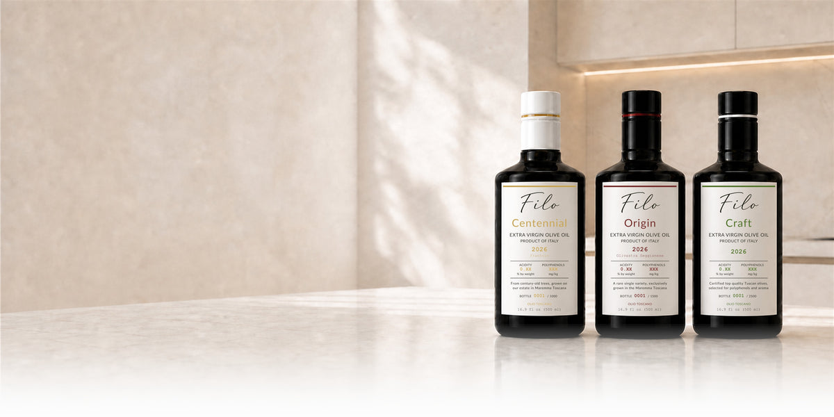 Three dark glass bottles of Filo extra virgin olive oil stand on a light marble surface in a warm, modern kitchen setting. The bottles are labeled Filo Centennial, Filo Origin, and Filo Craft, with white labels and gold and red accents.